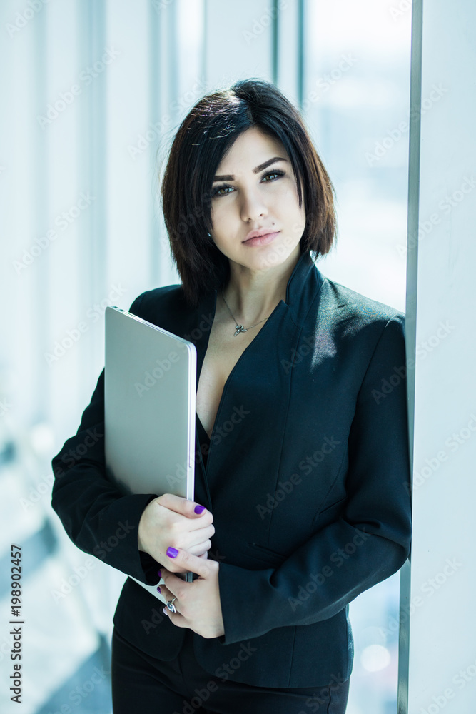 Young business woman working in her office holding closed laptop standing against panoramic window with a view on city Young business woman working in her office holding closed laptop standing against panoramic window with a view on city