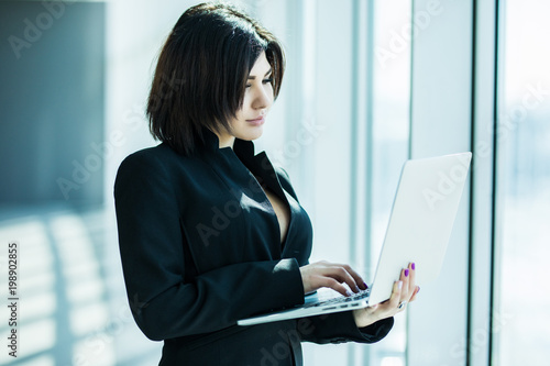 Portrait of young business woman working holding laptop standing against panoramic window with city view © dianagrytsku