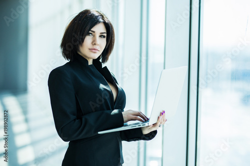 Portrait of young business woman working holding laptop standing against panoramic window with city view Free Download Portrait of young business woman working holding laptop standing against panoramic window with city view - GFXLoad