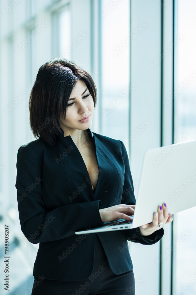 Businesswoman standing against office window holding laptop. Businesswoman in bright office. Businesswoman standing against office window holding laptop. Businesswoman in bright office.