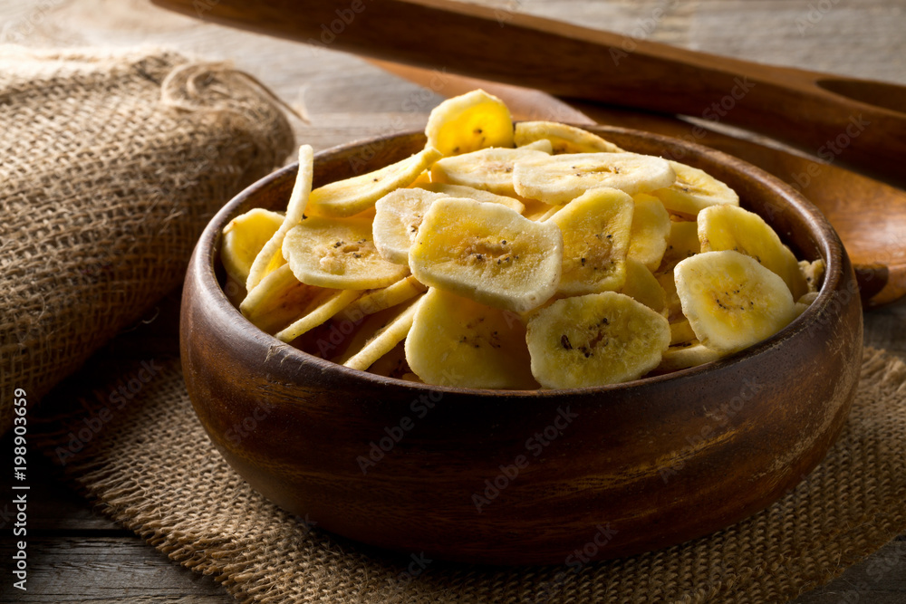Heap of dried banana chips snack in wooden bowl on dark table Heap of dried banana chips snack in wooden bowl on dark table