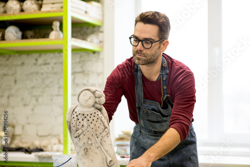 Obraz na plátně Portrait of Ceramist Dressed in an Apron Working on Clay Sculpture in Bright Ceramic Workshop
