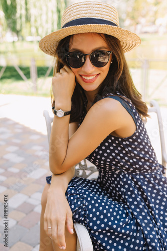 Portrait of charming style woman in the summer park wearing summer hat and black sunglasses and cute dress. She is looking at camera with wonderful smile. Background summer park. Summer holiday.