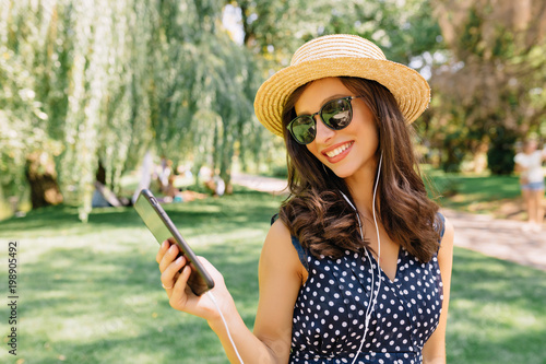 Photo of style woman is walking in the summer park wearing summer hat and black sunglasses and cute dress. She is listening music and dancing with great emotions. Background summer park. Holidays.