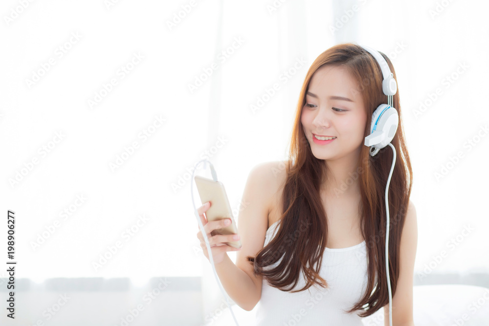 Naklejka premium Beautiful asian young woman enjoy listen music with headphone and holding smart mobile phone while sitting in bedroom, relax girl with earphone, leisure and technology concept.