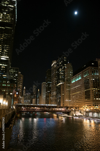 Chicago city riverwalk promenade at night with vintage drawbridge, illuminated urban downtown buildings and the moon. © Bruno Passigatti