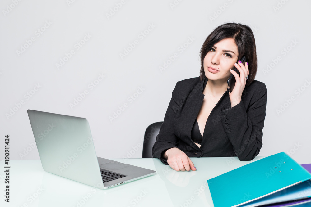 Portrait of a young and attractive professional business woman sitting at her office work desk having a telephone conversation and using a laptop computer, isolated on a white wall. Portrait of a young and attractive professional business woman sitting at her office work desk having a telephone conversation and using a laptop computer, isolated on a white wall.