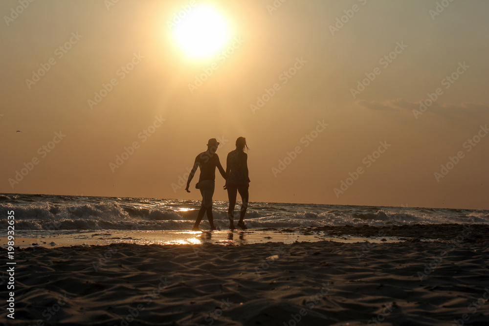 Young couple is walking in the water on summer beach. Sunset over the sea.Two silhouettes against the sun. Man and woman in holiday honeymoon trip.