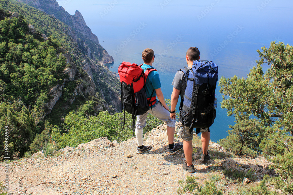 Two young guys with backpacks stand on a cliff above the sea