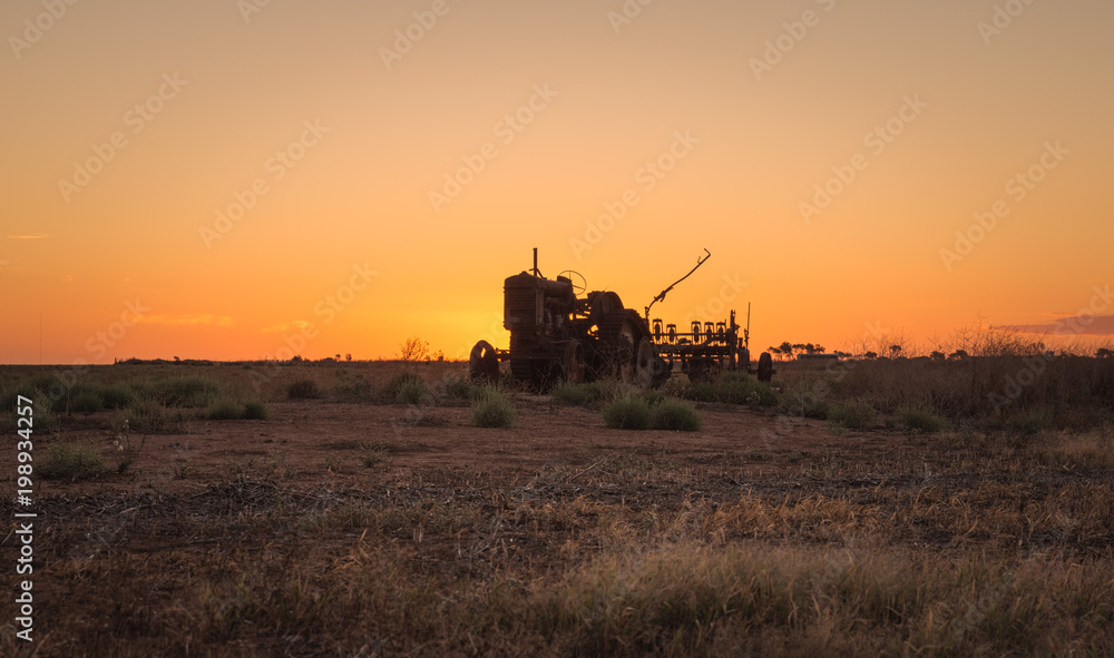 Old abandoned tractor in farm field with rusted agricultural equipment silhouetting at sunset