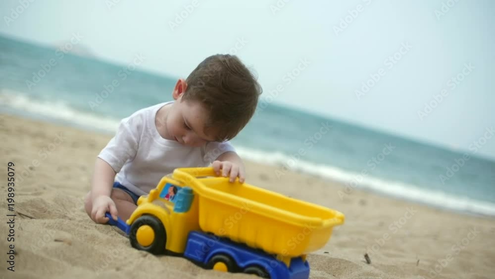 Two-year old beautiful baby toddler boy riding a toy cargo car on summer beach closeup. Slow motion Two-year old beautiful baby toddler boy riding a toy cargo car on summer beach closeup. Slow motion