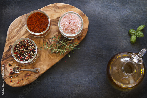 Close-up photo of colored dried spices in bowls on wooden board on black concreted table background with olive oil in jar