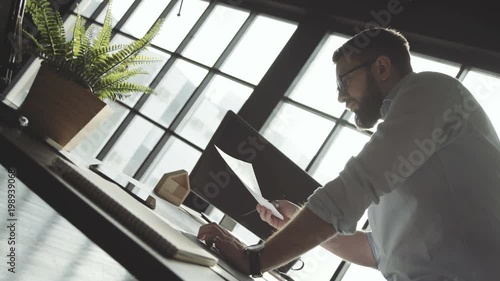 Wallpaper Mural Man with a beard in white shirt works against the background of a large panoramic window, leans on table. Young businessman works alone in bright spacious office, stylish accessories and plants. Torontodigital.ca
