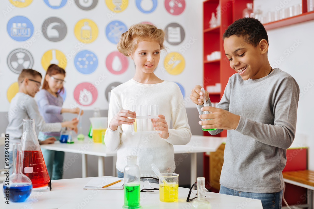 Interesting experiment. Joyful teenage boy adding green chemical to the flask with a pipette and smiling while his experiment partner looking at him with curiosity Interesting experiment. Joyful teenage boy adding green chemical to the flask with a pipette and smiling while his experiment partner looking at him with curiosity