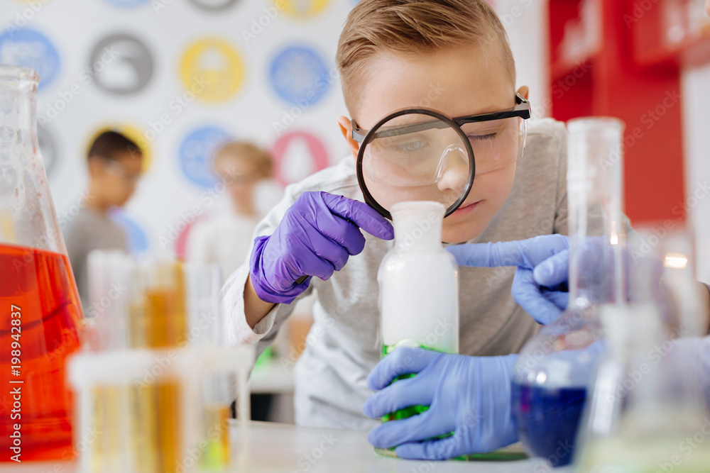 Careful examination. Pleasant teenage male student using a magnifying glass and examining a green substance in the flask during his chemistry class Careful examination. Pleasant teenage male student using a magnifying glass and examining a green substance in the flask during his chemistry class