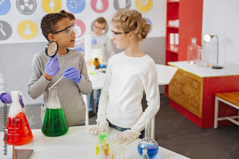Fresh ideas. Charming teenage classmates discussing chemistry experiment while the boy adding a few drops of chemical to the flask Fresh ideas. Charming teenage classmates discussing chemistry experiment while the boy adding a few drops of chemical to the flask