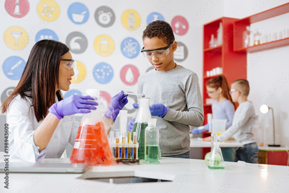 Vital help. Caring chemistry teacher adding agent to the flask of her student and helping him to conduct an experiment successfully Vital help. Caring chemistry teacher adding agent to the flask of her student and helping him to conduct an experiment successfully