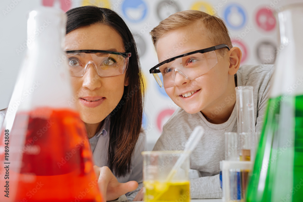 Favorite science. The close up of an upbeat teenage student and his young chemistry teacher watching the chemicals reacting in the flasks during the class in the lab Favorite science. The close up of an upbeat teenage student and his young chemistry teacher watching the chemicals reacting in the flasks during the class in the lab