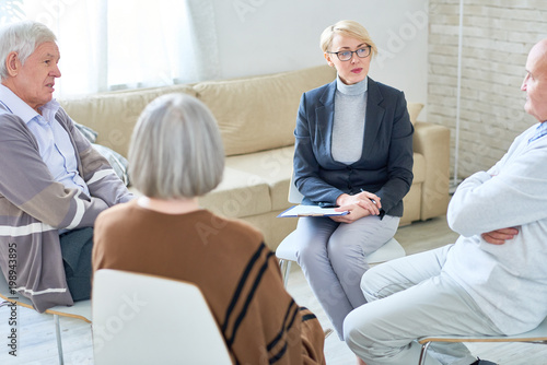 Portrait of blonde female psychiatrist wearing glasses leading group therapy session for senior people sitting in circle in retirement home, copy space