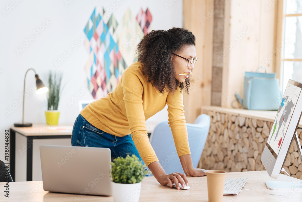 My new startup. Good-looking serious young curly-haired woman holding a pencil and working on her laptop while standing at her table My new startup. Good-looking serious young curly-haired woman holding a pencil and working on her laptop while standing at her table