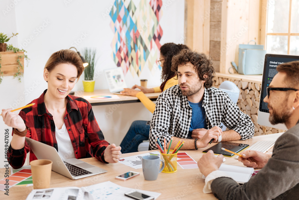 Obraz premium New ideas. Beautiful joyful blond woman smiling and showing information on her laptop and her colleagues sitting at the table with her