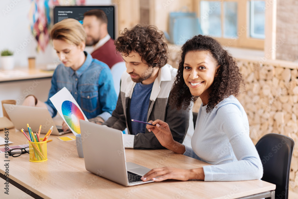 I feel great. Beautiful joyful curly-haired woman smiling and working on her laptop while sitting at the table with her colleagues I feel great. Beautiful joyful curly-haired woman smiling and working on her laptop while sitting at the table with her colleagues