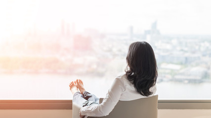 Life quality concept with business woman rear view relaxing sitting in rest on cozy armchair in modern hotel guest room or luxury home living room looking toward beautiful city urban scene
