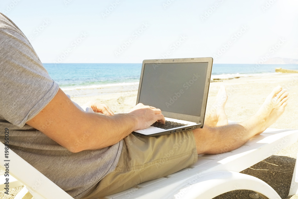 Fit young man using laptop sitting on white wood beach veranda porch. Freelance concept. Male programmer working coding on notebook computer, blank screen. Sunny, ocean sea shore. Copy space backgroun