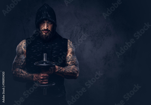 Portrait of a muscular bearded tattoed male wearing black sportswear with a hood, doing exercise with a dumbbell, isolated on a dark background.
