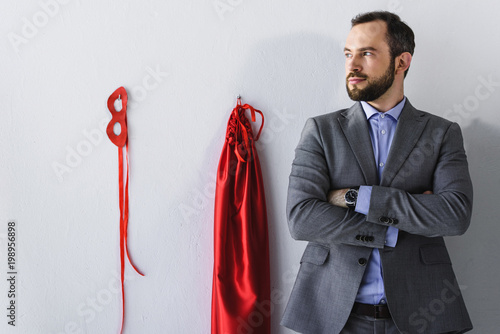 super businessman standing with crossed arms near mask and cape on wall in office