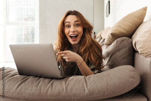 Image of happy european woman lying on sofa in living room, and pointing finger on screen of notebook with interest or surprise