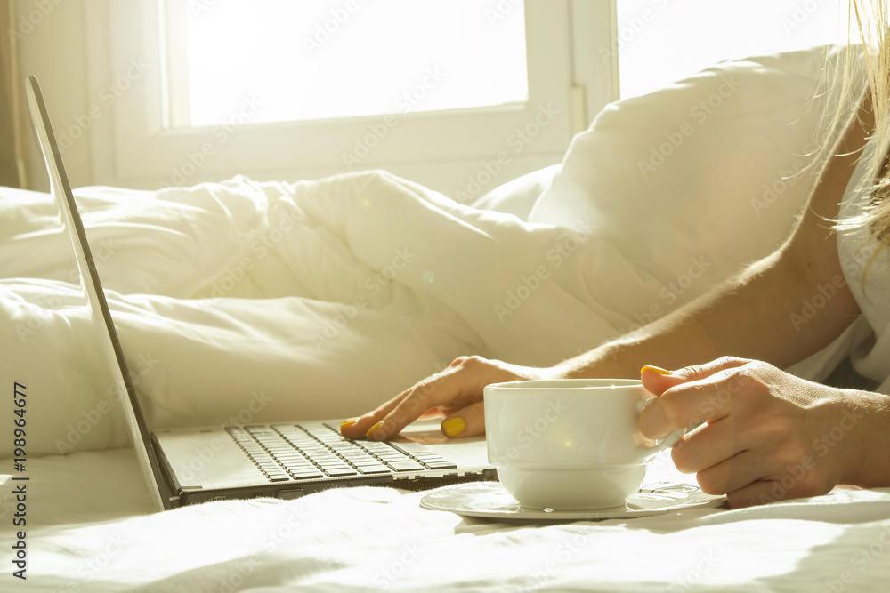 Close up, young blond woman in bed, white sheets linens holding cup of morning coffee, surfing browsing blogging eshopping on laptop. Perks of freelance concept. Blond female in tight shirt Background