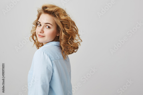 Studio portrait of good-looking slender woman with curly blonde hair turning and smiling at camera while stending in profile over gray background. Sensual girlfriend waits for her lover photo