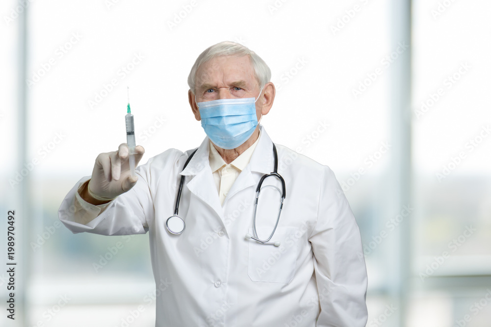 Old serious doctor holding syringe with vaccine. Ready to make injection. Bright blurred background. Old serious doctor holding syringe with vaccine. Ready to make injection. Bright blurred background.