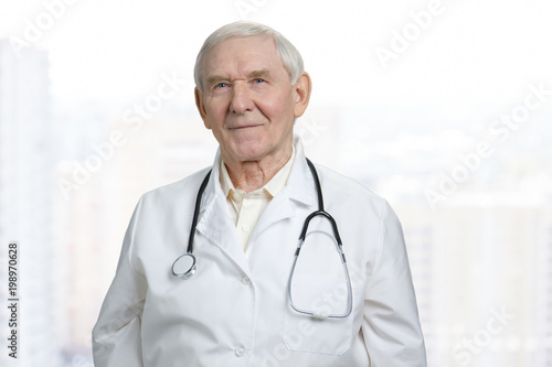 Portrait of smiling old caucasian doctor in lab coat. Senior physician in stethoscope in bright background.