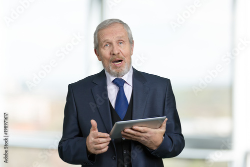 Speech of old businessman with tablet. Portrait of senior man with modern gadget is talking, bright blurred background.