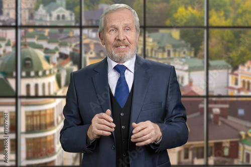 Old european politician man portrait. Senior man in suit talking, checkered windows background with view on small european town.