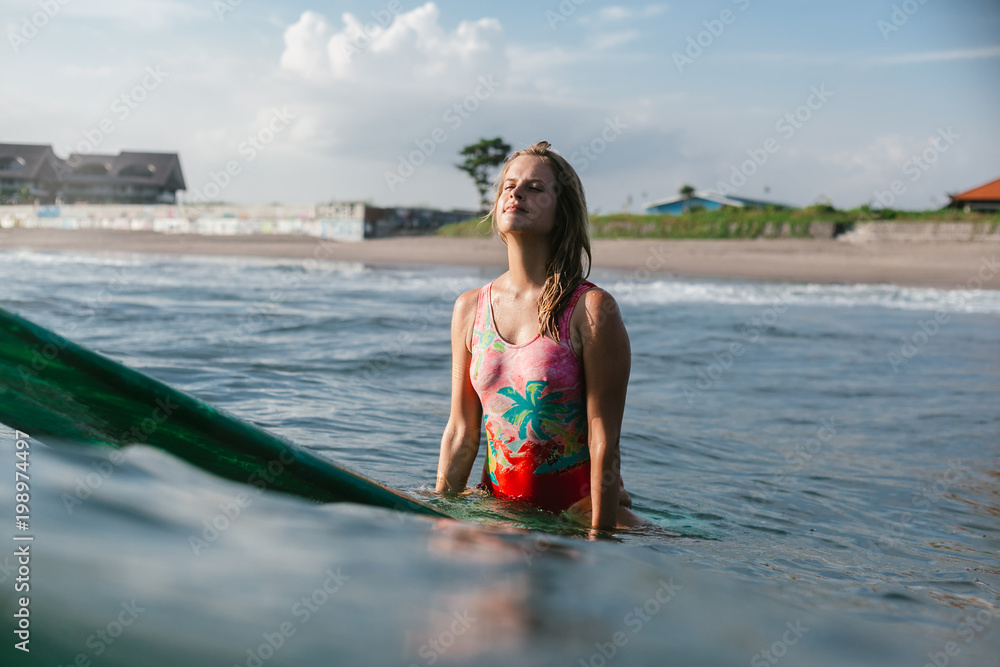 portrait of woman in swimming suit resting on surfing board in ocean with beach on background