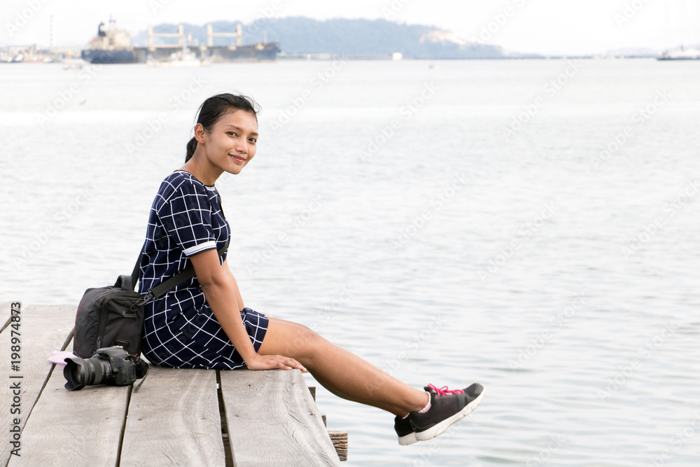 young woman with camera sitting on a wooden pier over the sea. Asian tourist relaxing by the sea, Penang, Malaysia.
