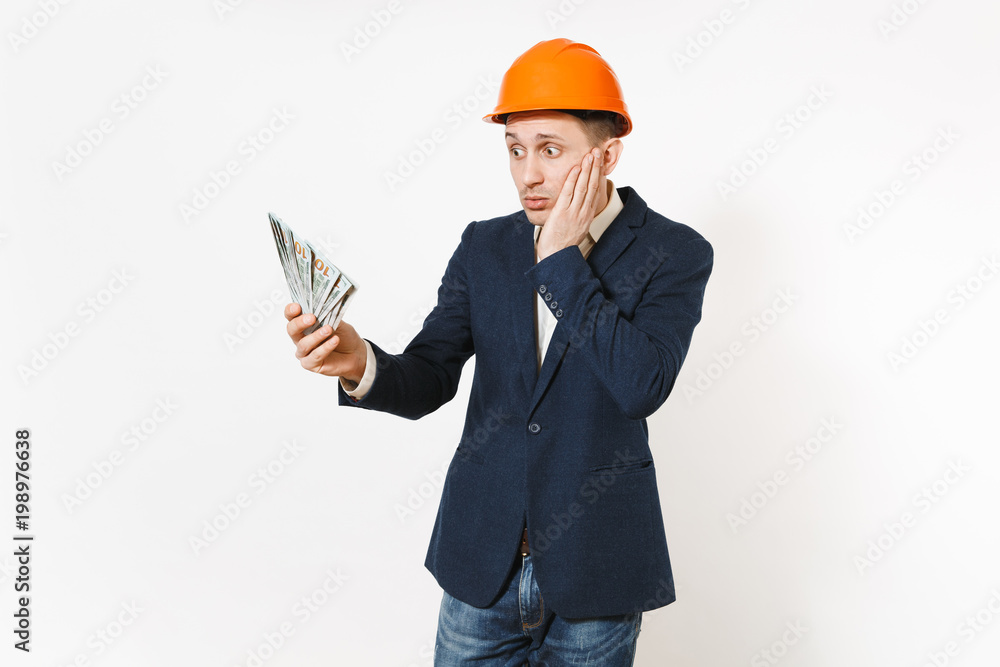 Young concerned businessman in dark suit, protective hardhat holding bundle of dollars, cash money and spreading hands isolated on white background. Male worker for advertisement. Business concept. Young concerned businessman in dark suit, protective hardhat holding bundle of dollars, cash money and spreading hands isolated on white background. Male worker for advertisement. Business concept.