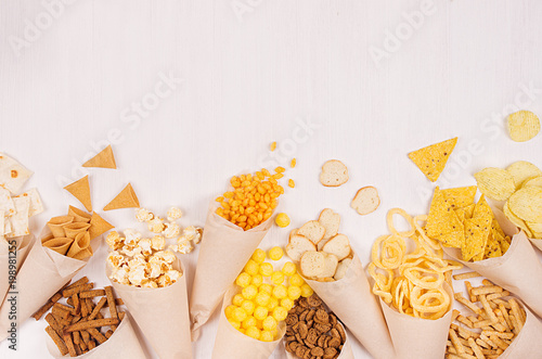 Beige paper cones with bright crunchy fast food snacks - nachos, popcorn, croutons, chips on white wood board, copy space. photo