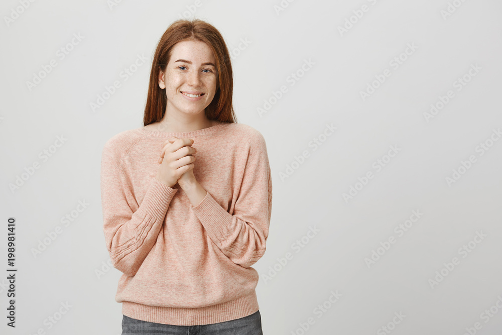 Lovely charming redhead european woman standing in tender pose, clasping hands near chest and smiling broadly over gray background. Girl is touched by romantic scene in movie Lovely charming redhead european woman standing in tender pose, clasping hands near chest and smiling broadly over gray background. Girl is touched by romantic scene in movie