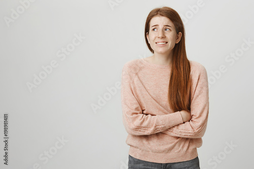 Indoor portrait of cute ginger girl standing with crossed hands on belly, feeling awkward or suffering from pain while looking aside, standing against gray background. Woman has stomachache