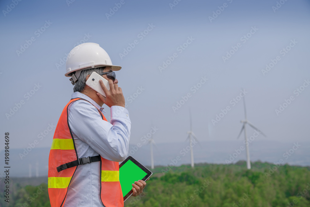 Senior engineer man with safety helmet talking abount working and holding tablet for checking condition of wind turbine on the high hill mountain.Wind turbine electric generator,clean energy concept. Senior engineer man with safety helmet talking abount working and holding tablet for checking condition of wind turbine on the high hill mountain.Wind turbine electric generator,clean energy concept.