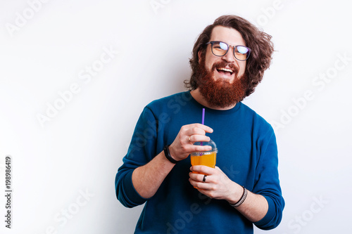 Cheerful bearded hipster man with long hair and eye glasses holding plastic cup of fresh juice, orange, vitamins