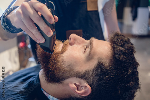 Side view close-up of the head of a redhead young man and the hand of a skilled barber, trimming his beard with an electric trimmer in a trendy hair salon for men