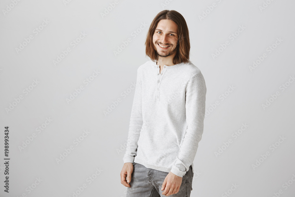 Modern prince in shiny white sweater. Portrait of attractive slender male with beard and long hair standing half-turned and smiling broadly, inviting cute woman on coffee, expressing friendliness Modern prince in shiny white sweater. Portrait of attractive slender male with beard and long hair standing half-turned and smiling broadly, inviting cute woman on coffee, expressing friendliness
