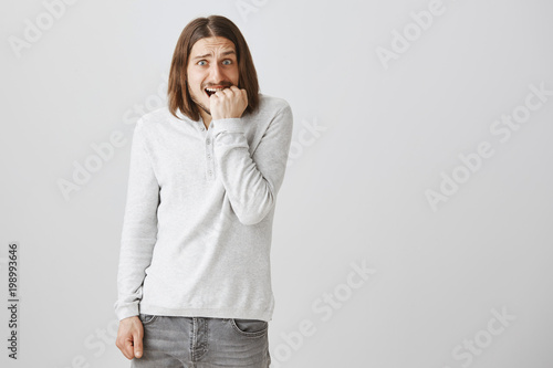 How should I fix my mistake. Portrait of worried ashamed adult male with long hair and beard shrugging, biting fist and being anxious and nervous, hiding secret or being guilty over gray background