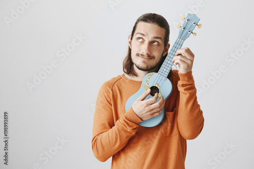 Tuning strings to show amazing playing skill. Handsome funny musician in orange sweater holding ukulele near face, pulling string while making faces and looking aside, fooling around over gray wall.