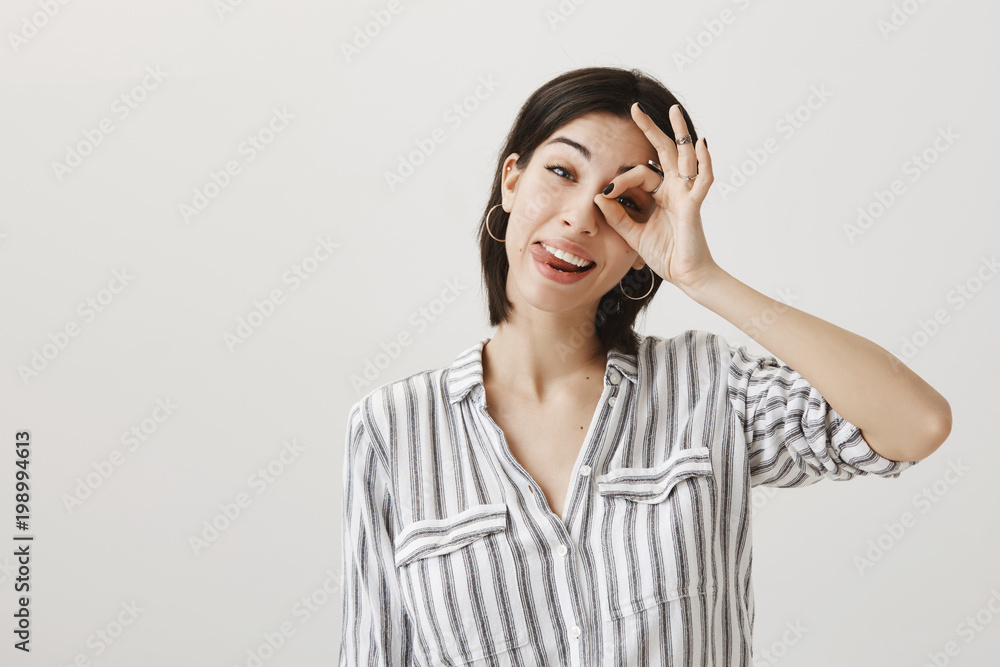 Indoor shot of playful attractive european female with short haircut and stylish outfit showing okay or fine gesture over eye, sticking out tongue and making faces being in good mood over gray wall Indoor shot of playful attractive european female with short haircut and stylish outfit showing okay or fine gesture over eye, sticking out tongue and making faces being in good mood over gray wall